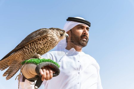 Téléchargez les photos : Homme du Moyen-Orient portant un kandura arabe émirati traditionnel dans le désert et tenant un oiseau de faucon - Arabe musulman adulte dans les dunes de sable de Dubaï - en image libre de droit