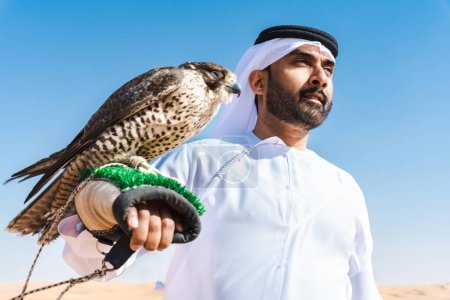 Téléchargez les photos : Homme du Moyen-Orient portant un kandura arabe émirati traditionnel dans le désert et tenant un oiseau de faucon - Arabe musulman adulte dans les dunes de sable de Dubaï - en image libre de droit