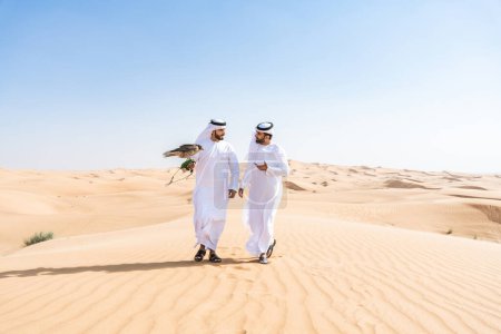Téléchargez les photos : Deux hommes du Moyen-Orient portant des kandura arabes emirati traditionnels se lient dans le désert et tenant un oiseau de faucon - des amis musulmans arabes se rencontrent dans les dunes de sable de Dubaï - en image libre de droit