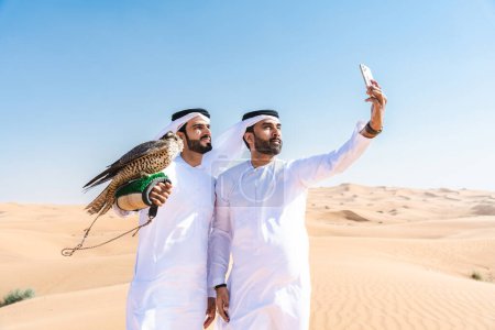 Photo for Two middle-eastern men wearing traditional emirati arab kandura bonding in the desert and holding a falcon bird - Arabian muslim friends meeting at the sand dunes in Dubai - Royalty Free Image