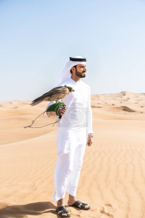 Foto de Hombre de Oriente Medio vestido con el tradicional emirati árabe kandura en el desierto y sosteniendo un pájaro halcón árabe musulmán adulto en las dunas de arena en Dubai - Imagen libre de derechos