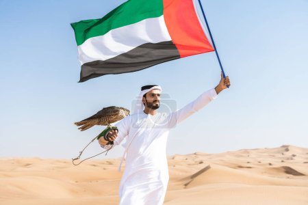 Foto de Hombre de Oriente Medio con el tradicional emirati kandura árabe en el desierto y sosteniendo un pájaro halcón y la bandera de los Emiratos Árabes Unidos - persona musulmana adulta en las dunas de arena en Dubai - Imagen libre de derechos