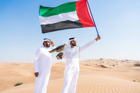 Foto de Dos hombres de Oriente Medio que llevan puesto el tradicional emirati árabe kandura vinculación en el desierto y la celebración de un pájaro halcón y la bandera de los Emiratos Árabes Unidos amigos musulmanes se reúnen en las dunas de arena en Dubai - Imagen libre de derechos