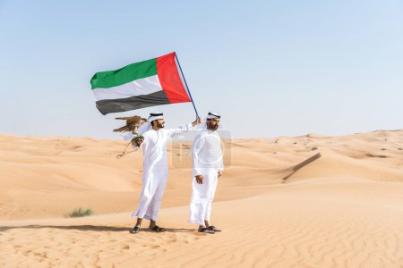 Téléchargez les photos : Deux hommes du Moyen-Orient portant des kandouras arabes emirati traditionnelles se lient dans le désert et tenant un oiseau de faucon et le drapeau des Émirats arabes unis - amis musulmans arabes réunis dans les dunes de sable à Dubaï - en image libre de droit