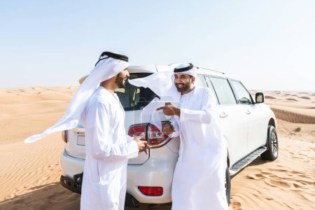 Photo for Two middle-eastern men wearing traditional emirati arab kandura driving a 4x4 car in the desert - Arabian muslim friends meeting at the sand dunes in Dubai for an excursion - Royalty Free Image