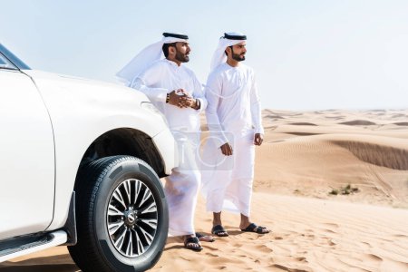 Foto de Dos hombres de Oriente Medio que llevan el tradicional emirati árabe kandura conduciendo un coche 4x4 en el desierto - amigos musulmanes árabes que se reúnen en las dunas de arena en Dubai para una excursión - Imagen libre de derechos