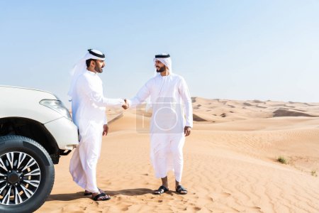 Téléchargez les photos : Deux hommes du Moyen-Orient portant une kandura arabe emirati traditionnelle conduisant une voiture 4x4 dans le désert - des amis musulmans arabes se réunissant dans les dunes de sable de Dubaï pour une excursion - en image libre de droit