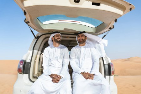 Téléchargez les photos : Deux hommes du Moyen-Orient portant une kandura arabe emirati traditionnelle conduisant une voiture 4x4 dans le désert - des amis musulmans arabes se réunissant dans les dunes de sable de Dubaï pour une excursion - en image libre de droit