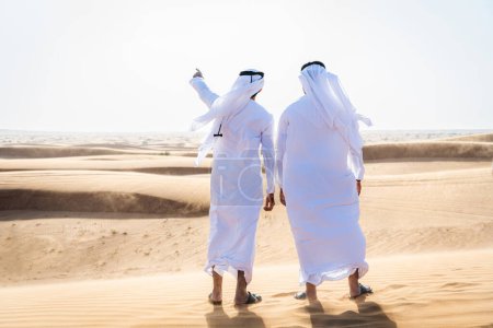 Foto de Dos hombres de Oriente Medio que llevan puesto el tradicional emirati árabe kandura vinculación en el desierto - amigos musulmanes árabes se reúnen en las dunas de arena en Dubai - Imagen libre de derechos