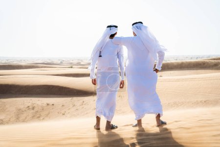 Téléchargez les photos : Deux hommes du Moyen-Orient portant des liens traditionnels de kandura arabe emirati dans le désert - des amis musulmans arabes se réunissant dans les dunes de sable de Dubaï - en image libre de droit
