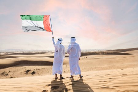 Foto de Dos hombres de oriente medio que llevan puesto el tradicional emirati árabe kandura que se une en el desierto y enarbolan la bandera de los Emiratos Árabes Unidos para celebrar el día nacional - amigos musulmanes árabes que se reúnen en las dunas de arena en Dubai - Imagen libre de derechos