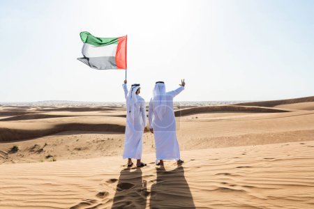 Photo for Two middle-eastern men wearing traditional emirati arab kandura bonding in the desert and holiding the UAE flag to celebrate national day - Arabian muslim friends meeting at the sand dunes in Dubai - Royalty Free Image