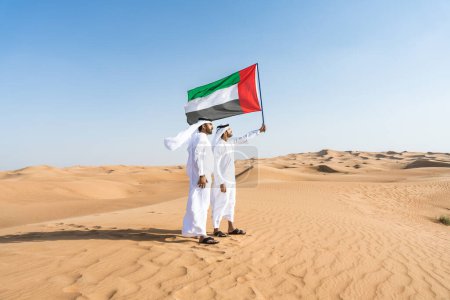 Foto de Dos hombres de oriente medio que llevan puesto el tradicional emirati árabe kandura que se une en el desierto y enarbolan la bandera de los Emiratos Árabes Unidos para celebrar el día nacional - amigos musulmanes árabes que se reúnen en las dunas de arena en Dubai - Imagen libre de derechos