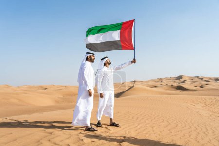 Téléchargez les photos : Deux hommes du Moyen-Orient portant le traditionnel kandura arabe emirati se lient dans le désert et planant le drapeau des Émirats arabes unis pour célébrer la fête nationale - des amis musulmans arabes se rencontrent dans les dunes de sable de Dubaï - en image libre de droit