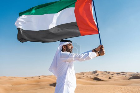 Téléchargez les photos : Homme du Moyen-Orient vêtu de kandura arabe émirati traditionnelle dans le désert tenant le drapeau des EAU - Arabe musulman adulte dans les dunes de sable de Dubaï célébrant le patriotisme le jour de la fête nationale - en image libre de droit