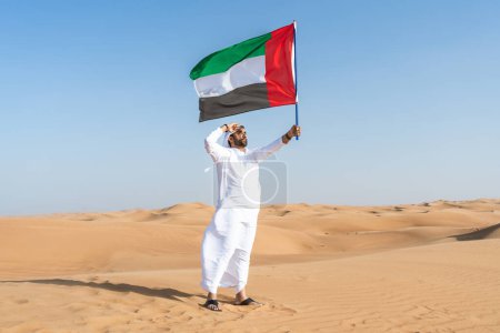 Foto de Hombre de Oriente Medio que viste el tradicional emirati kandura árabe en el desierto sosteniendo la bandera de los Emiratos Árabes Unidos - árabe musulmán adulto en las dunas de arena en Dubai celebrando el patriotismo en el día nacional - Imagen libre de derechos