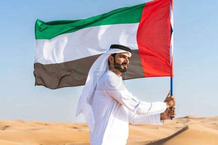Téléchargez les photos : Homme du Moyen-Orient vêtu de kandura arabe émirati traditionnelle dans le désert tenant le drapeau des EAU - Arabe musulman adulte dans les dunes de sable de Dubaï célébrant le patriotisme le jour de la fête nationale - en image libre de droit