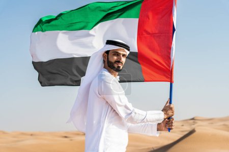 Foto de Hombre de Oriente Medio que viste el tradicional emirati kandura árabe en el desierto sosteniendo la bandera de los Emiratos Árabes Unidos - árabe musulmán adulto en las dunas de arena en Dubai celebrando el patriotismo en el día nacional - Imagen libre de derechos