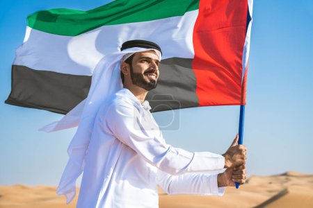 Photo for Middle-eastern man wearing traditional emirati arab kandura  in the desert holding the UAE flag - Arabian muslim adult person at the sand dunes in Dubai celebrating patriotism on national day - Royalty Free Image