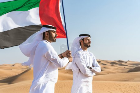 Photo for Two middle-eastern men wearing traditional emirati arab kandura bonding in the desert and holiding the UAE flag to celebrate national day - Arabian muslim friends meeting at the sand dunes in Dubai - Royalty Free Image