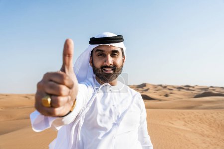 Téléchargez les photos : Beau et réussi homme du Moyen-Orient portant kandura arabe traditionnel emirati dans le désert - Arabe musulman adulte dans les dunes de sable à Dubaï - en image libre de droit