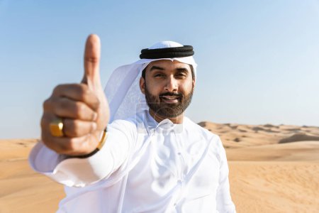 Téléchargez les photos : Beau et réussi homme du Moyen-Orient portant kandura arabe traditionnel emirati dans le désert - Arabe musulman adulte dans les dunes de sable à Dubaï - en image libre de droit