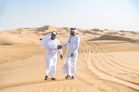 Photo for Two middle-eastern men wearing traditional emirati arab kandura bonding in the desert - Arabian muslim friends meeting at the sand dunes in Dubai - Royalty Free Image