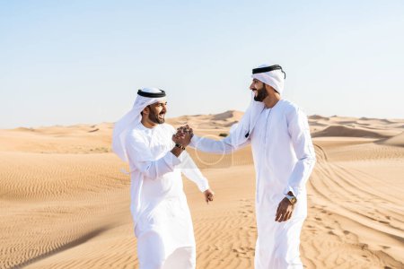 Foto de Dos hombres de Oriente Medio que llevan puesto el tradicional emirati árabe kandura vinculación en el desierto - amigos musulmanes árabes se reúnen en las dunas de arena en Dubai - Imagen libre de derechos