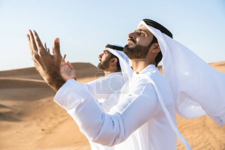 Téléchargez les photos : Deux hommes du Moyen-Orient portant des liens traditionnels de kandura arabe emirati dans le désert - des amis musulmans arabes se réunissant dans les dunes de sable de Dubaï - en image libre de droit