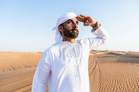 Foto de Hombre guapo y exitoso de Oriente Medio que usa el tradicional emirati árabe kandura en el desierto - árabe musulmán adulto en las dunas de arena en Dubai - Imagen libre de derechos