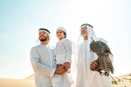 Téléchargez les photos : Famille de trois générations faisant un safari dans le désert de Dubaï. Grand-père, fils et petit-fils passer du temps ensemble dans la nature et former leur oiseau de faucon. - en image libre de droit