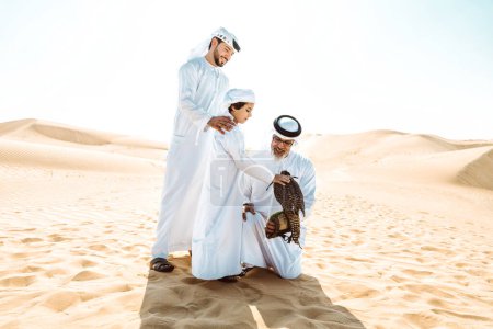 Téléchargez les photos : Famille de trois générations faisant un safari dans le désert de Dubaï. Grand-père, fils et petit-fils passer du temps ensemble dans la nature et former leur oiseau de faucon. - en image libre de droit