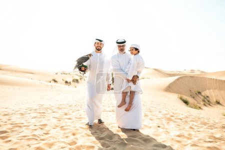 Foto de Familia de tres generaciones haciendo un safari en el desierto de Dubai. Abuelo, hijo y nieto pasando tiempo juntos en la naturaleza y entrenando a su ave halcón. - Imagen libre de derechos