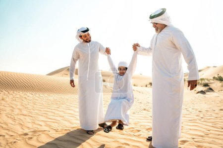 Photo for Three generation family making a safari in the desert of Dubai wearing white kandura outfit. Grandfather, son and grandson spending time together in the nature. - Royalty Free Image