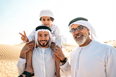 Téléchargez les photos : Famille de trois générations faisant un safari dans le désert de Dubaï en tenue blanche kandura. Grand-père, fils et petit-fils passer du temps ensemble dans la nature. - en image libre de droit