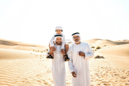 Téléchargez les photos : Famille de trois générations faisant un safari dans le désert de Dubaï en tenue blanche kandura. Grand-père, fils et petit-fils passer du temps ensemble dans la nature. - en image libre de droit