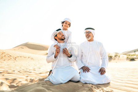 Photo for Three generation family making a safari in the desert of Dubai wearing white kandura outfit. Grandfather, son and grandson spending time together in the nature. - Royalty Free Image