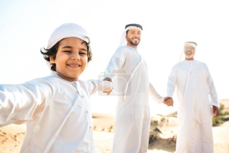 Foto de Familia de tres generaciones haciendo un safari en el desierto de Dubai vistiendo traje de kandura blanco. Abuelo, hijo y nieto pasando tiempo juntos en la naturaleza. - Imagen libre de derechos
