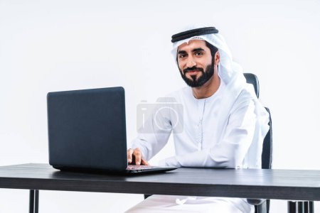 Téléchargez les photos : Emirate homme d'affaires du Moyen-Orient portant kandora travaillant dans le bureau, Dubaï Beau homme d'affaires d'entreprise assis au bureau de l'ordinateur et travaillant à distance avec un ordinateur portable - en image libre de droit