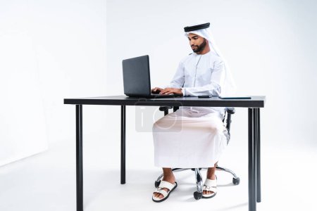 Foto de Emirate hombre de negocios de Oriente Medio con kandora trabajando en la oficina, Dubai - Hombre de negocios corporativo guapo sentado en el escritorio de la computadora y el trabajo remoto con el ordenador portátil - Imagen libre de derechos