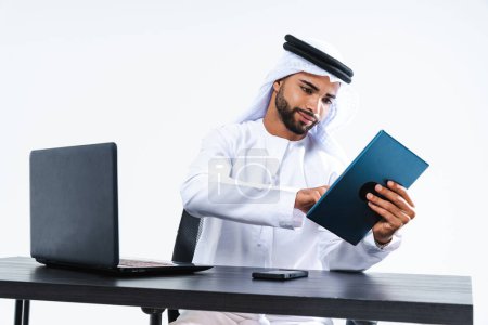 Foto de Emirate hombre de negocios de Oriente Medio con kandora trabajando en la oficina, Dubai - Hombre de negocios corporativo guapo sentado en el escritorio de la computadora y el trabajo remoto con el ordenador portátil - Imagen libre de derechos