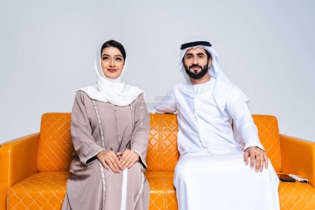 Photo for Beautiful arab middle-eastern happy couple of lovers wearing traditional abaya and kandora sitting on sofa at home- Arabic muslim adult people bonding and having fun in Dubai, United Arab Emirates - Royalty Free Image
