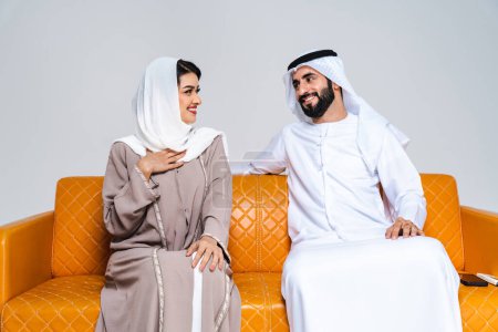 Foto de Hermosa pareja de amantes del Oriente Medio feliz árabe vistiendo abaya tradicional y kandora sentado en el sofá en casa musulmanes árabes adultos vinculación y divertirse en Dubai, Emiratos Árabes Unidos - Imagen libre de derechos