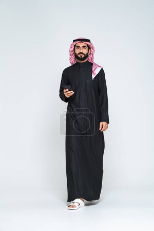 Foto de Hombre árabe árabe saudí de Oriente Medio guapo con ropa tradicional saudí en el estudio - hombre de negocios árabe musulmán adulto con retrato thwab aislado sobre fondo gris - Imagen libre de derechos