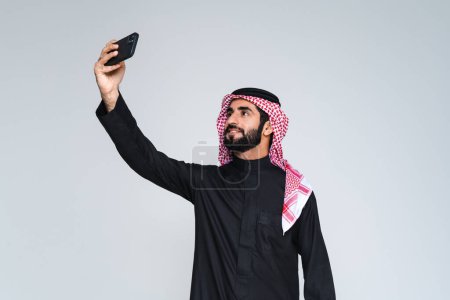 Foto de Hombre árabe árabe saudí de Oriente Medio guapo con ropa tradicional saudí en el estudio - hombre de negocios árabe musulmán adulto con retrato thwab aislado sobre fondo gris - Imagen libre de derechos