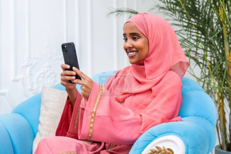 Foto de Mujer feliz de Oriente Medio vistiendo abaya en casa - Hermoso retrato musulmán adulto femenino en el apartamento, conceptos sobre la vida doméstica y el estilo de vida islámico - Imagen libre de derechos