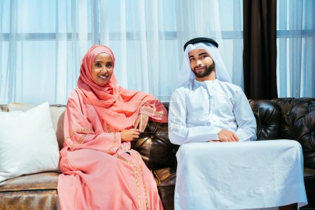 Foto de Feliz pareja de oriente medio que usa ropa árabe tradicional en casa: marido y mujer árabes casados que se unen en el apartamento, conceptos sobre la relación, la vida doméstica y el estilo de vida emirati. - Imagen libre de derechos