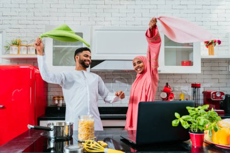 Foto de Feliz pareja de oriente medio que usa ropa árabe tradicional en casa: marido y mujer árabes casados que se unen en el apartamento, conceptos sobre la relación, la vida doméstica y el estilo de vida emirati. - Imagen libre de derechos