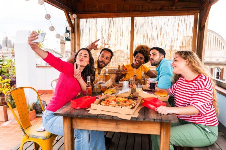 Foto de Grupo multiétnico de jóvenes amigos felices que cenan barbacoa en la azotea en casa: jóvenes adultos alegres y multiétnicos que se divierten y se unen en un balcón con terraza con vistas a la ciudad, comer y beber. - Imagen libre de derechos