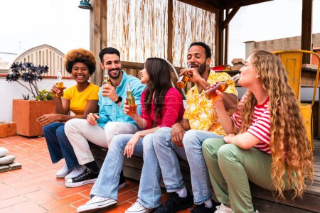 Foto de Grupo multiétnico de jóvenes amigos felices que cenan barbacoa en la azotea en casa: jóvenes adultos alegres y multiétnicos que se divierten y se unen en un balcón con terraza con vistas a la ciudad, comer y beber. - Imagen libre de derechos